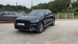 Тёмно‑синий Genesis GV70 2021 года, общий вид спереди‑сбоку на стоянке дилера; видна радиаторная решётка, колёса и фон с забором.