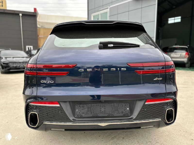 Задняя часть Genesis GV70: фонарьная группа, надпись GENESIS и двойной выхлоп на фоне ангаров и ограждения стоянки.