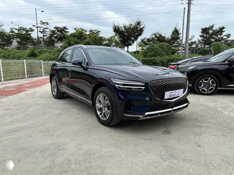 Под углом спереди‑сбоку показан тёмно‑синий Genesis GV70 на асфальтовой площадке с другими машинами и зеленью на заднем плане.