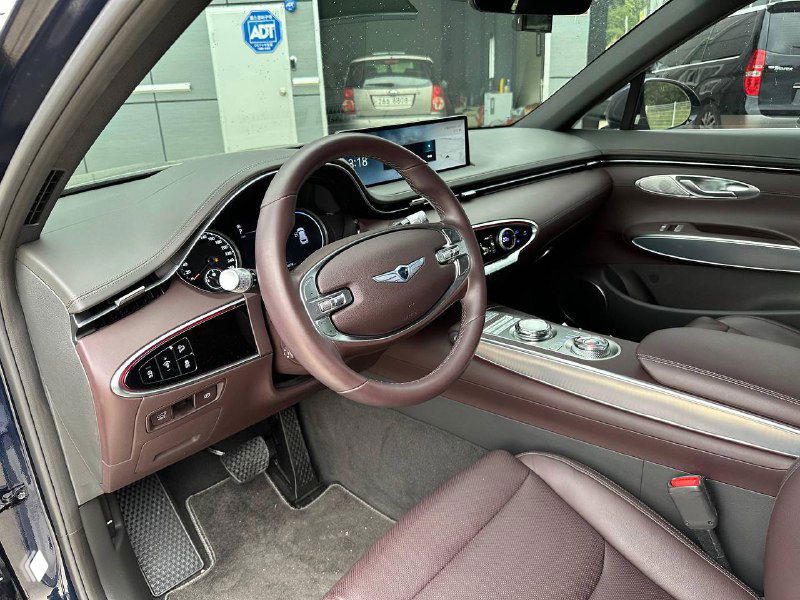 Водительское место Genesis GV70: рулевое колесо, щиток приборов и часть сиденья в коричневой кожаной отделке, видны кнопки управления.