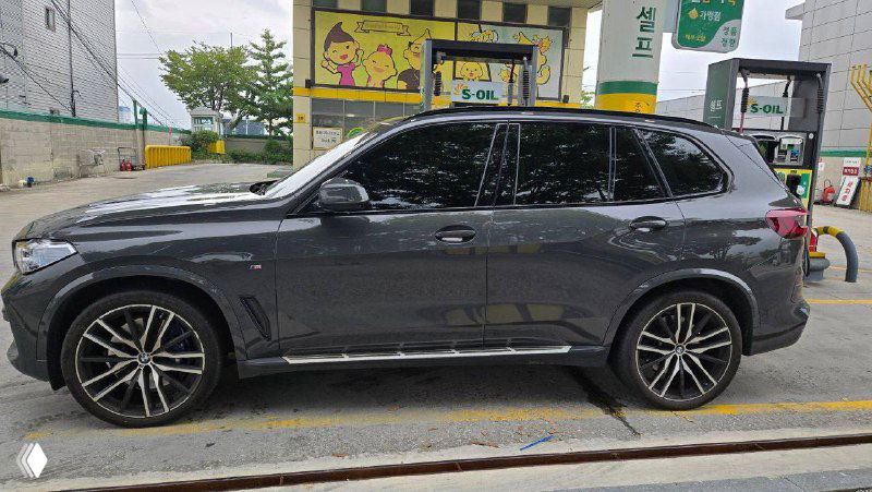 Боковой профиль BMW X5 40D 2022, серый металлик, крупный план колёс и линий кузова, автомобиль на парковке у заправки.