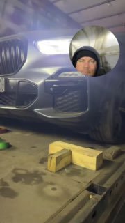 Крупный план передней части BMW с горящими фарами в тёплом гаражном свете; видны доски и инструменты рядом с автомобилем.