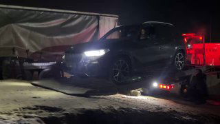 Автомобиль на рампе эвакуатора в тёмное время, машина частично затемнена, видны рабочие фонари и платформа грузовика.