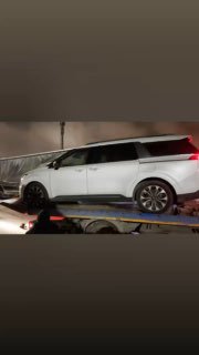 Ещё один ракурс белого минивэна на эвакуаторе; снимок вертикальной ориентации показывает транспортировку автомобиля на площадке в тёмное время.