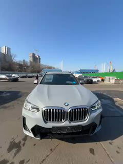 Белый BMW X4 20i xDrive M Sport, вид спереди на стоянке таможни Владивостока; на лобовом стекле видны документы и бирки.