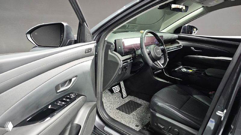 Передняя часть салона Hyundai Tucson: водительское сиденье, дверная карта с элементами управления и текстильные коврики на фоне открытой двери.