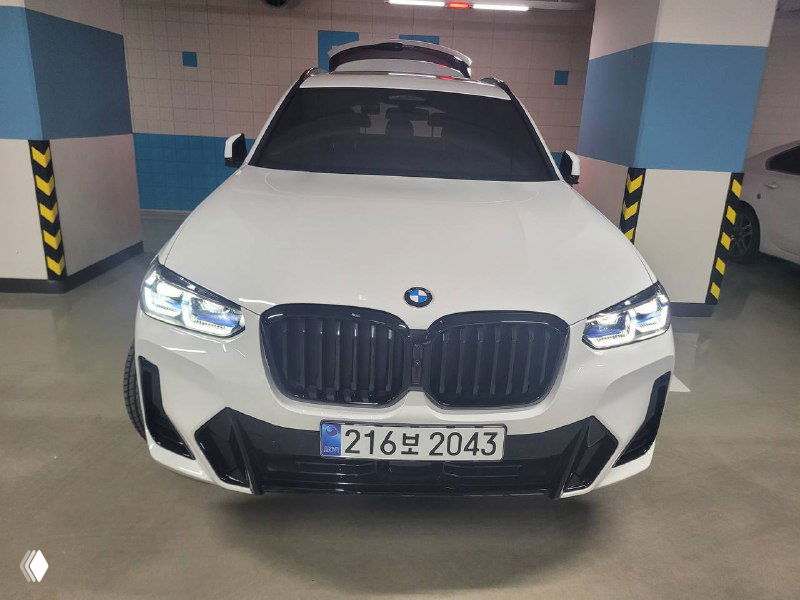 Фронтальный вид белого BMW X3 xDrive 20i MSport в паркинге, подчёркнутые решётка радиатора и светодиодные фары на фоне стен парковки.