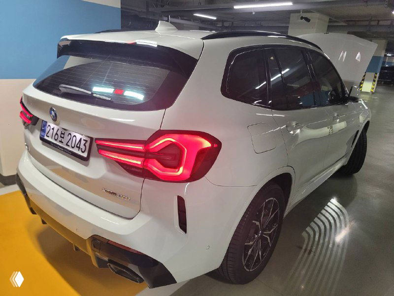 Задний левый ракурс BMW X3 xDrive 20i MSport белого цвета, подсвеченные задние фонари и вид на экстерьер сзади в паркинге.