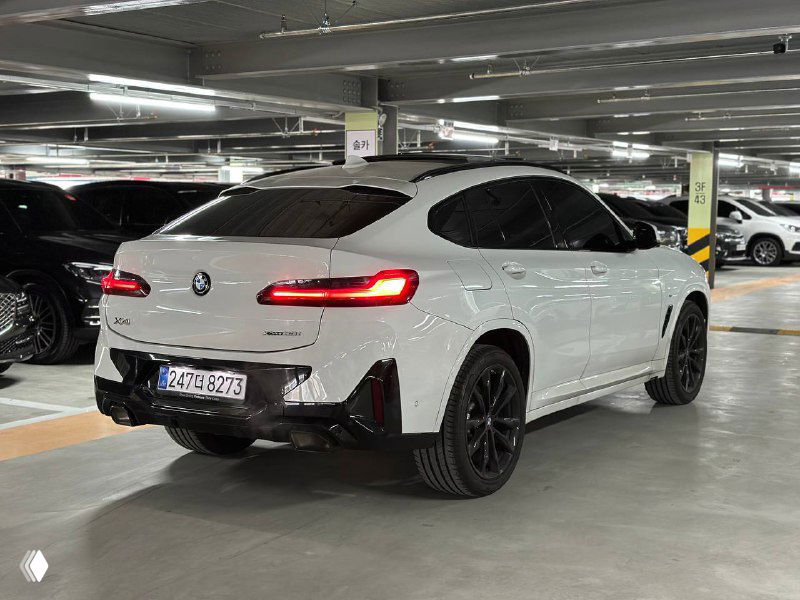 Вид сзади на BMW X4 в белом цвете — включены задние фонари, видна форма багажника и чёрные элементы диффузора на парковке.