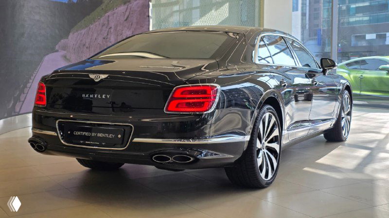 Задний ракурс черного Bentley Flying Spur в салоне дилера: видны фирменные задние фонари, две пары патрубков выхлопа и шильдик модели на багажнике.