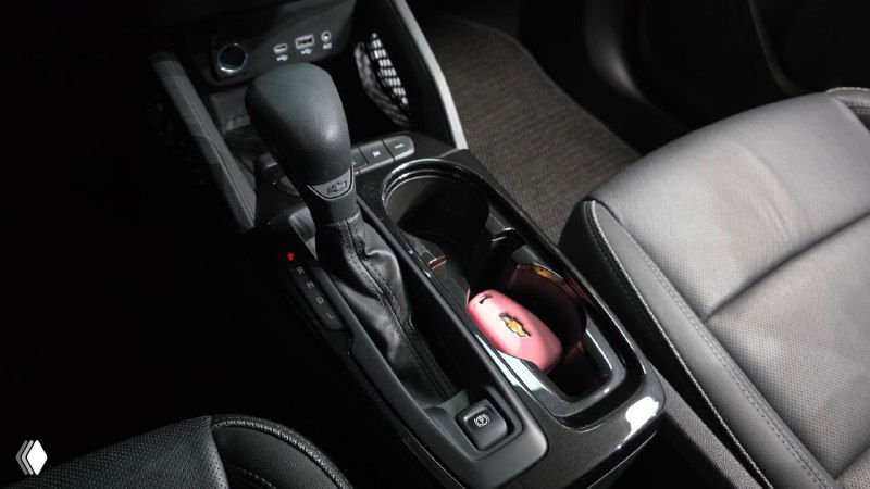 Фрагмент центральной консоли и селектора коробки передач Chevrolet Trailblazer, видны подстаканники и отделение с мелочами, чёрная отделка.