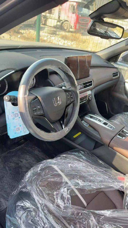 Водительская зона Honda UR-V: руль, крупный сенсорный дисплей мультимедиа и элементы управления на руле, сиденье в защитной плёнке, вертикальная ориентация снимка.