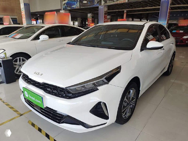 Белый Kia K3, вид в три четверти спереди со стороны водителя в салоне автосалона; показаны легкосплавные диски и профиль кузова автомобиля.