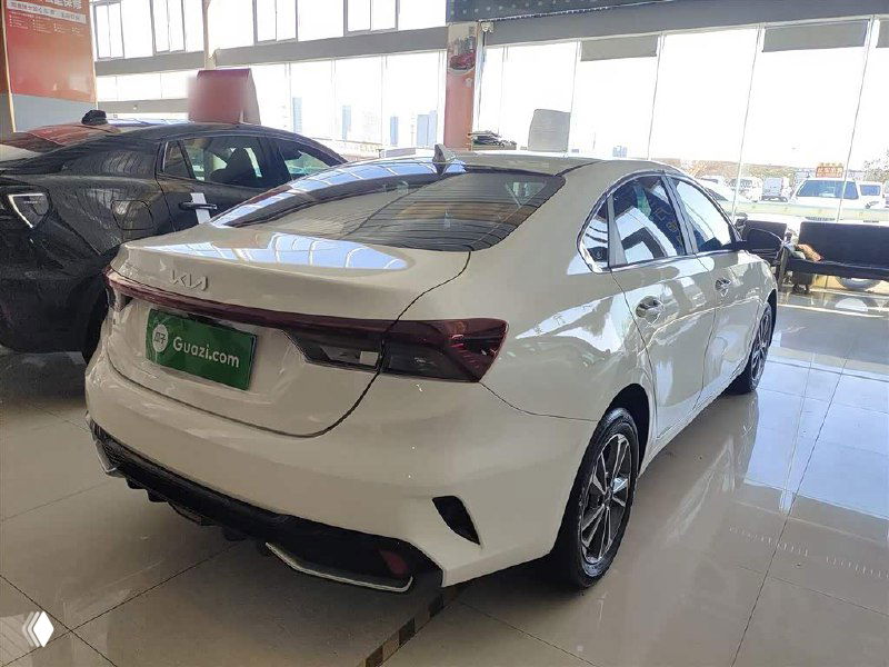 Задняя часть белого Kia K3 в выставочном зале, видно оптику, бампер и общий силуэт седана при дневном свете сквозь витрину.