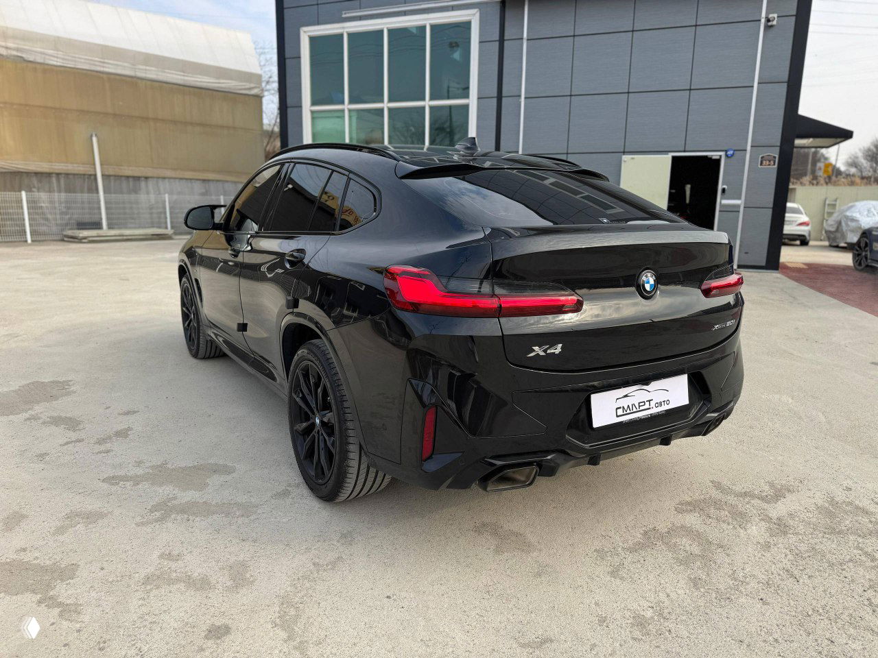 Задний вид BMW X4 20i XDrive MSport в открытом паркинге, заметны стопы и эмблема модельного ряда на крышке багажника.