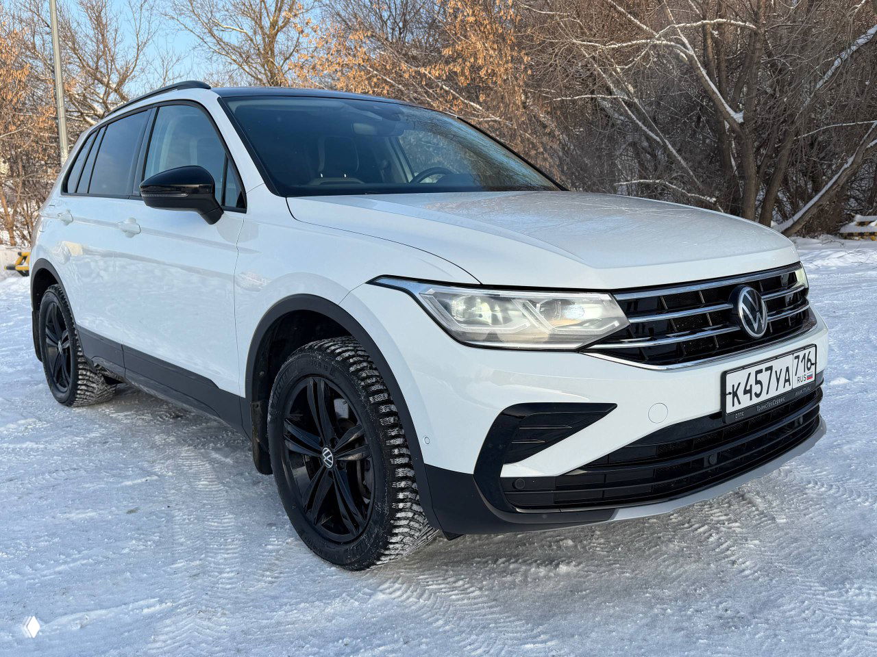 Volkswagen Tiguan Urban Sport 2022, боковой передний ракурс, белый кузов с чёрной крышей и зимние шины на снегу у деревьев.