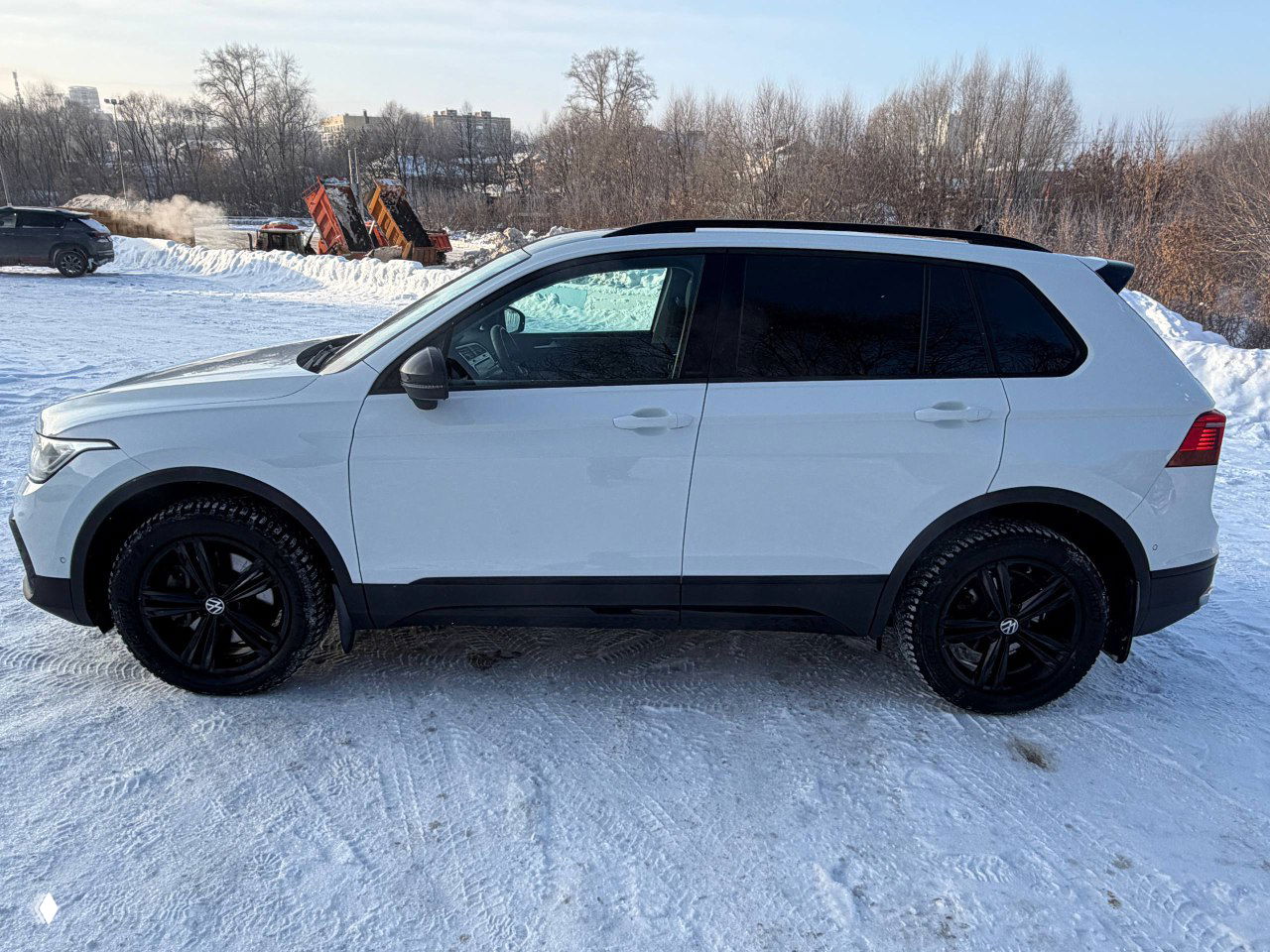 Боковой вид Volkswagen Tiguan 2022: белый кузов, контрастные чёрные диски, зимняя стоянка и городской фон на заднем плане.