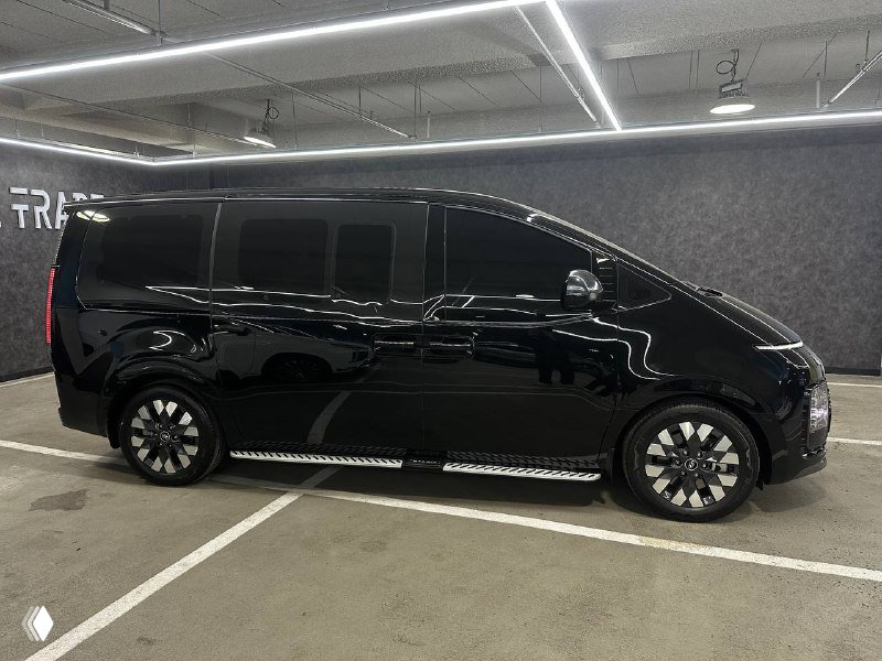 Черный Hyundai Staria, вид сбоку на стоянке, показаны дверные линии, колёсные диски и глянцевая поверхность кузова под потолочным светом.