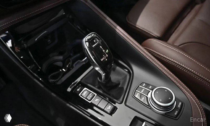 Селектор коробки передач и элементы управления мультимедиа BMW X1; видна центральная консоль и элементы управления в окружении коричневой обивки.