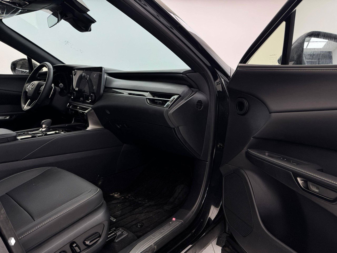 Панорамный вид салона Lexus RX300 2025 через открытую переднюю дверь — торпедо, экран и интерьер в чёрной отделке.