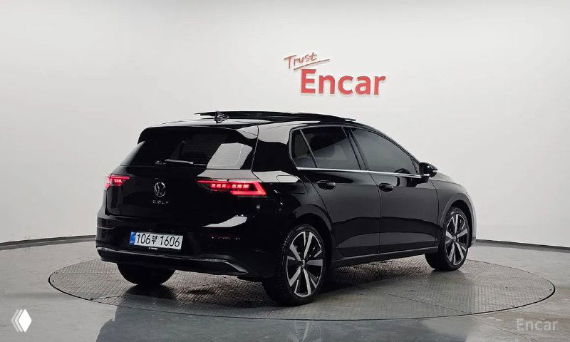 Задний три четверти чёрного Volkswagen Golf 2025 года в салоне, видно задние фонари и открытую багажную область издалека.