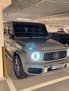 Угол обзора на Mercedes G63 AMG в паркинге: акцент на решётку радиатора, фары и часть кузова серебристого цвета с отражениями.