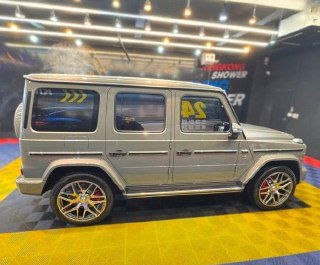 Боковой профиль Mercedes G63 AMG на выставочной подложке: серебристый кузов, крупные колёсные диски и оформление стен шоурума с подсветкой.