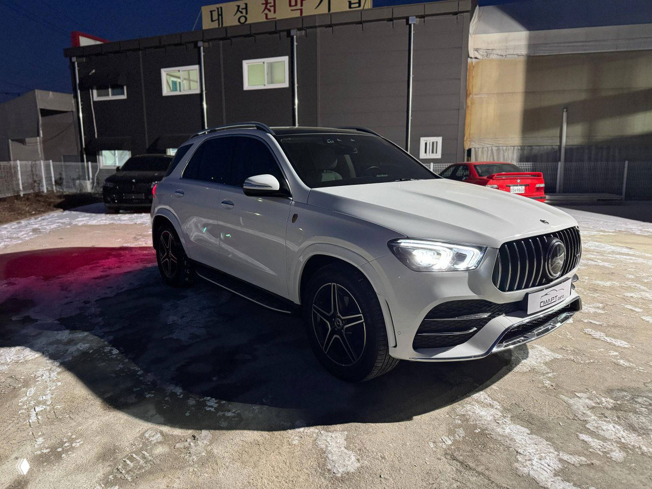 Белый Mercedes GLE 300D в вечернем освещении, машина на открытой стоянке, видна крыша и боковые двери.