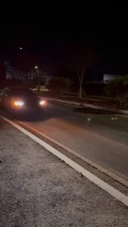 Тёмный автомобиль на дорожной платформах ночью с включёнными фарами — автомобиль ожидает погрузки и отправки из Кореи в РФ