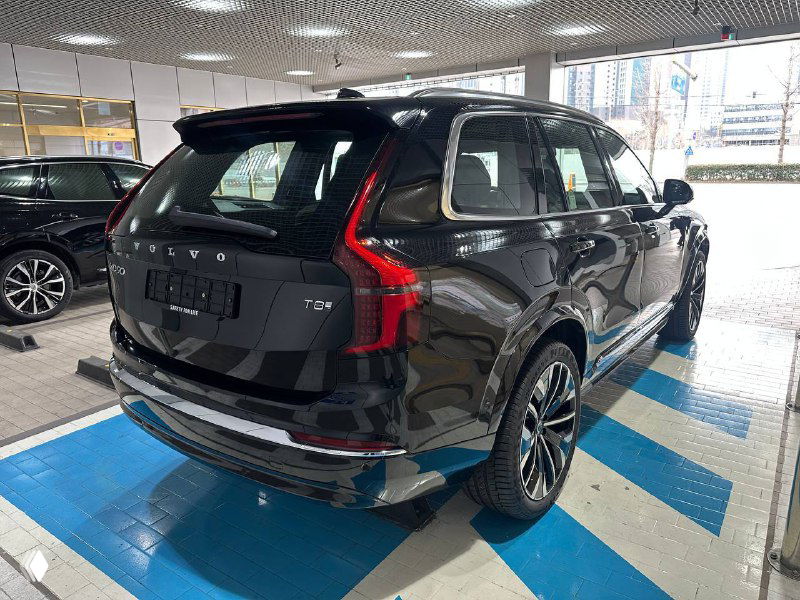 Volvo XC90 T8 2025, задняя три четверти, чёрный кузов, характерные вертикальные фонари и багажная часть в дилерском зале.