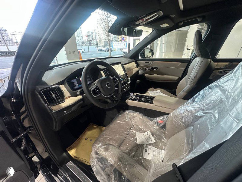 Передняя часть салона Volvo XC90 T8: панель приборов, большой мультимедийный экран и водительское кресло под защитной плёнкой в дилерских условиях.