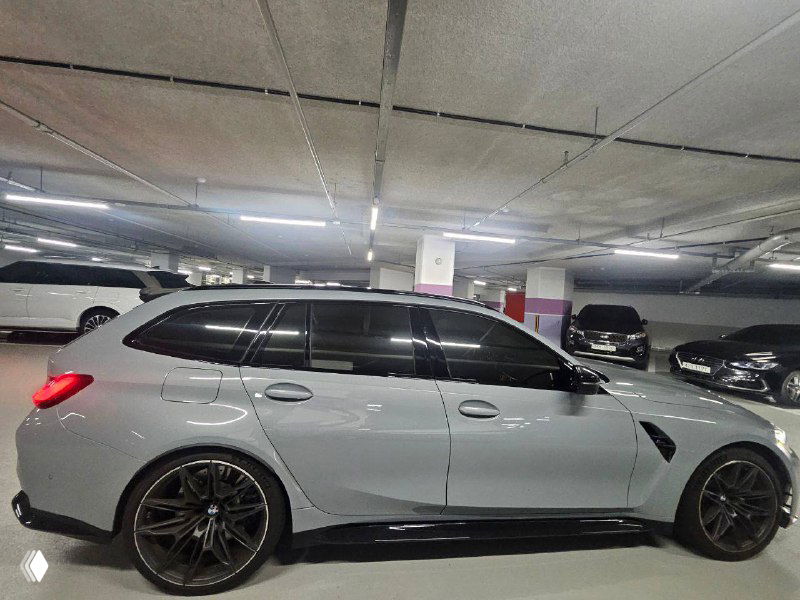 Вид с заднего левого бокового ракурса BMW M3 Touring в подземной стоянке, затемнённые стекла и спортивные элементы кузова на фоне других машин.