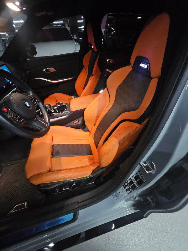 Передние сиденья BMW M3 с яркой оранжевой обивкой и вставками из алькантары, чётко видна спортивная форма кресел и боковая поддержка.