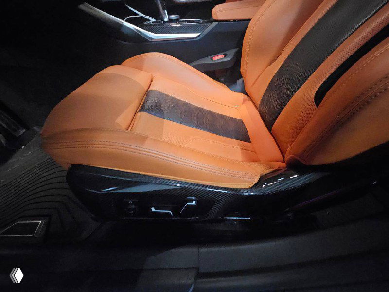 Крупный план переднего сиденья BMW M3: регулировки, боковая поддержка и детали отделки на фоне оранжевой кожи и карбоновых элементов.