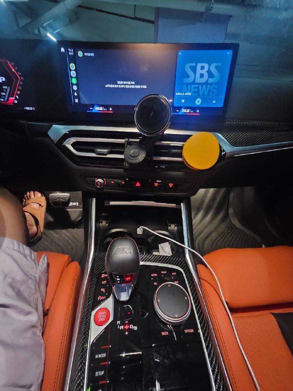 Центральная консоль BMW M3 с мультимедийным экраном, селектором передач, управлением климатом и подключёнными зарядными кабелями.