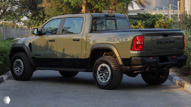Вид сзади Dodge Ram Rho 2025 цвета Canyon Lake — профиль грузового отсека, задние фары и эмблема RHO, автомобиль на улице рядом с ограждением.