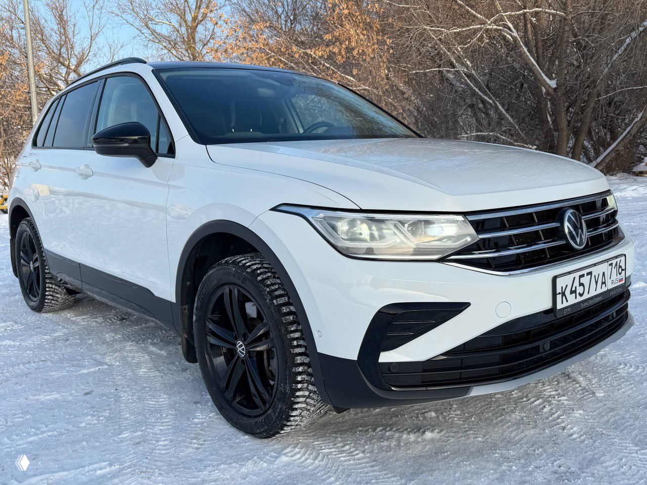 Трёхчетвертной ракурс белого Volkswagen Tiguan Urban Sport 2022 с чёрными дисками и зимней резиной на зимней стоянке, видна боковая часть и фары.