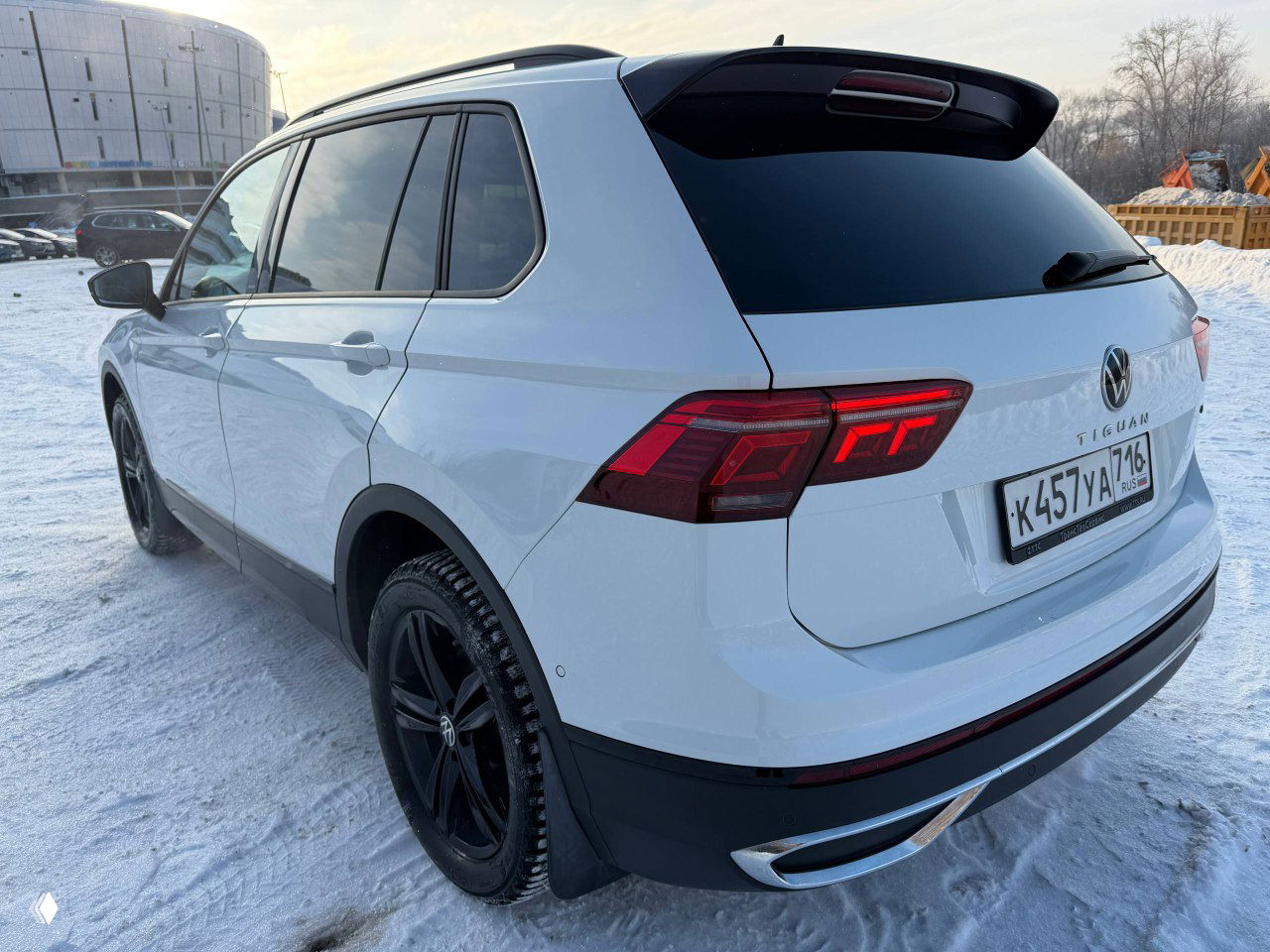 Вид сзади-угла белого Volkswagen Tiguan Urban Sport 2022, видны задние фары, брызговики и чёрные диски; автомобиль снят на снежной площадке при дневном свете.