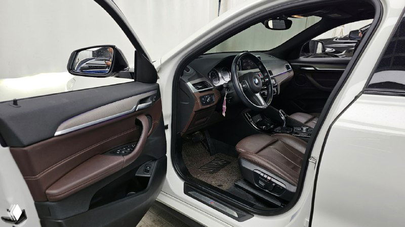 Открытая водительская дверь BMW X2: вид на переднее сиденье и внутреннюю панель двери, коричневый кожаный салон и элементы отделки.
