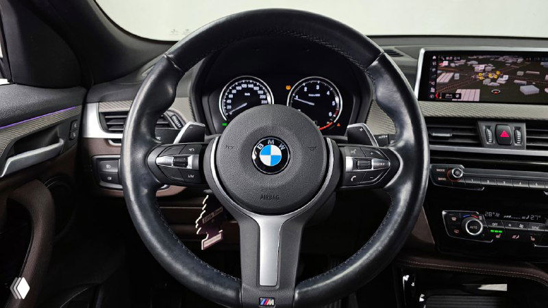 Руль и приборная панель BMW X2: крупный план руля с логотипом BMW и цифровыми приборами, элементы управления на руле видны чётко.
