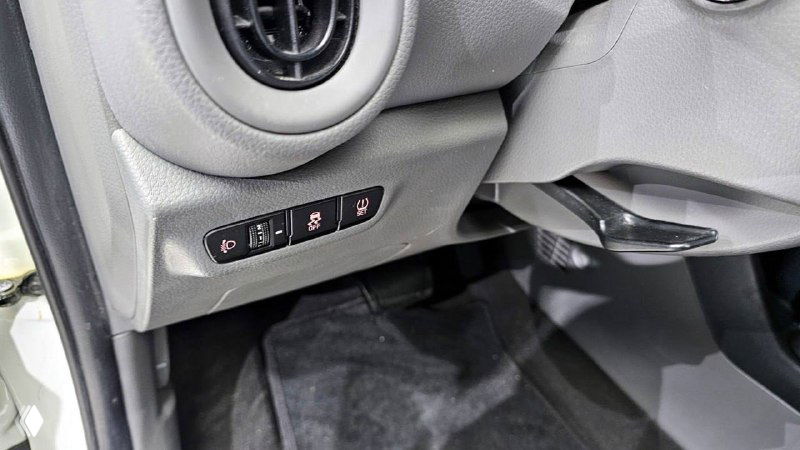 Детальный план нижней части панели водителя с блоком кнопок и воздуховодом, чёрный коврик и часть порога, демонстрация мелких деталей салона.