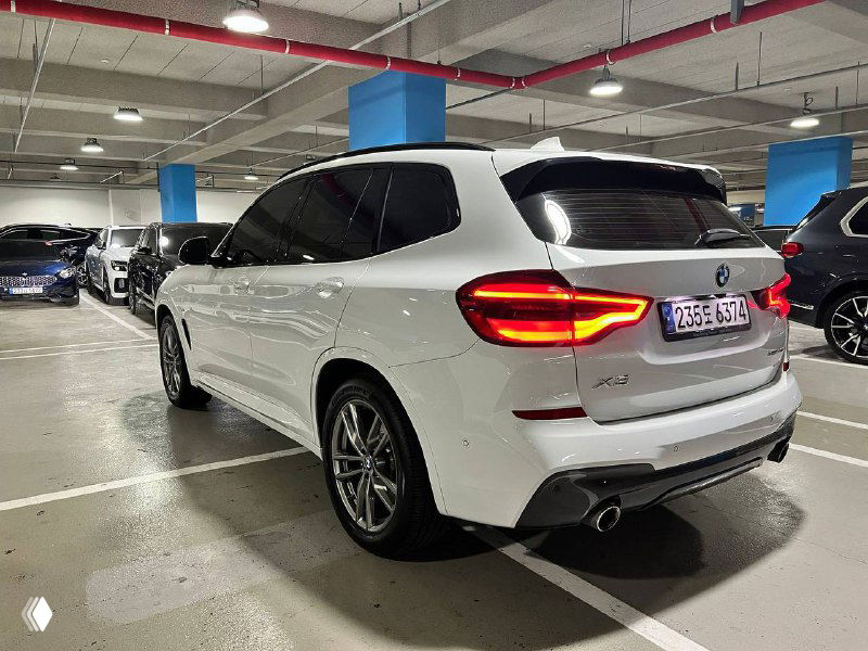 Задняя часть белого BMW X3 xDrive 20i M Sport, парковочный ракурс показывает фонари, выхлоп и общий профиль автомобиля в гараже.