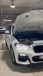 Белый BMW X3 с открытым капотом в подземной парковке, фронтальный ракурс показывает моторный отсек и состояние передней части автомобиля.