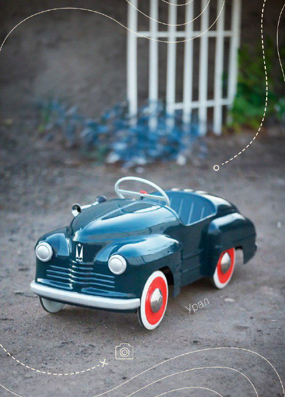 Тёмно‑синяя педальная ретро‑машина с округлым кузовом и белыми колёсами, фото в уличной обстановке демонстрирует классический дизайн игрушки.