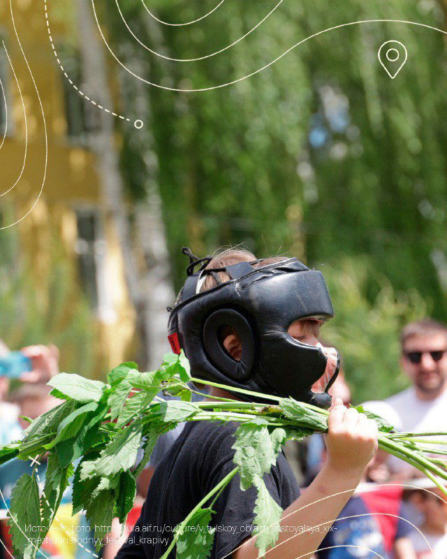 Участник в защитном шлеме держит ветки крапивы на фоне зрителей и деревьев — кадр крапивного боя на народном празднике в селе Крапивна.