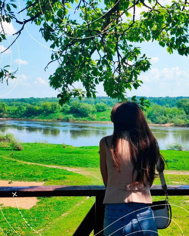 Вид на реку и зелёный берег из Тарусы: девушка стоит на набережной и смотрит на водную гладь и дальние леса в яркий летний день.