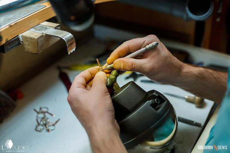 Ювелир аккуратно работает мелким инструментом с маленькой деталью украшения на прижимном держателе; виден рабочий стол и инструменты.