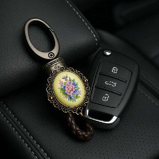 Ключи от автомобиля с брелоком-финифтью, брелок с миниатюрной цветочной росписью лежит на чёрной кожаной поверхности сиденья или подлокотника.