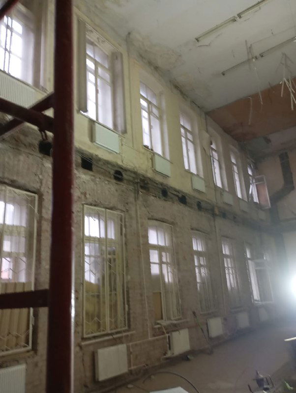 Двухъярусные окна и высокие стены в помещении на стадии реконструкции, видны повреждения штукатурки и подвешенные элементы потолка.