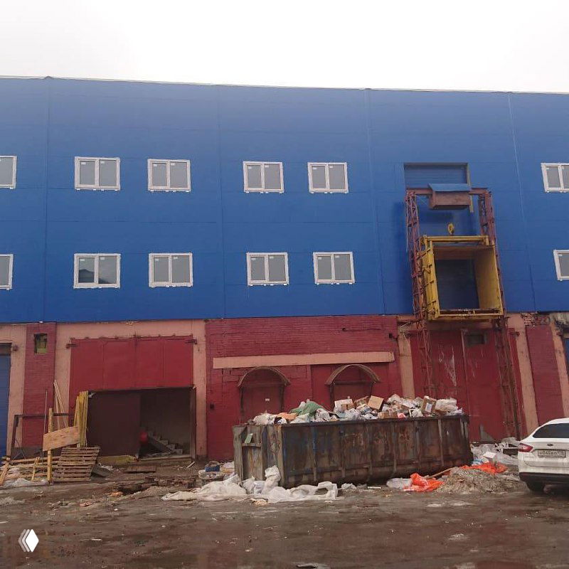 Строительная площадка у основания реконструированного корпуса: контейнеры для мусора, строительные материалы и техника на заднем плане у фасада нового здания.